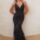 A woman stands against a textured, light-colored wall, wearing a form-fitting, floor-length black dress with floral lace detailing. The KENDRA PO2455 dress has a plunging neckline and thin straps. The sandy ground complements the light wall, creating a soft, neutral background. Angels Formal Wear