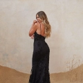 A woman with long, wavy hair stands on a sandy surface, facing away from the camera. She is wearing a floor-length, backless, black lace gown with spaghetti straps, designed by KENDRA PO2455. The background is a textured, light-colored wall with some sparse dried grass visible in the sand. Angels Formal Wear