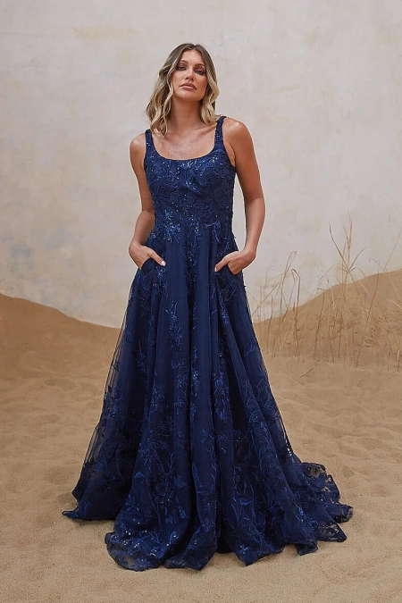 A woman stands on a sandy surface, wearing a long, sleeveless, deep blue gown adorned with floral embroidery. She has her hands in the dress's pockets and gazes slightly upward, reminiscent of LIR PO2471 against a light background behind her. Angels Formal Wear