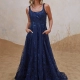 A woman stands on a sandy surface, wearing a long, sleeveless, deep blue gown adorned with floral embroidery. She has her hands in the dress's pockets and gazes slightly upward, reminiscent of LIR PO2471 against a light background behind her. Angels Formal Wear