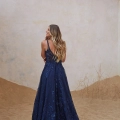 A woman with long, wavy hair stands with her back to the camera, wearing a dark blue, sleeveless gown with intricate detailing. The setting appears to be a sandy, partially indoor environment with a simple, textured wall in the background and "LIR PO2471" subtly embossed near the hem. Angels Formal Wear