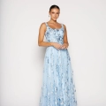 A woman stands against a plain background, gracefully donning a floor-length, sleeveless light blue gown adorned with delicate floral patterns. She has her hair styled back and poses with her hands gently clasped in front of her, embodying the timeless elegance of the LIR PO2471. Angels Formal Wear