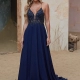 A woman stands on a sandy floor wearing the DAYLA PO2458 deep blue evening gown with a beaded, lace bodice and spaghetti straps. Her long hair is styled in loose waves, and she poses against a rustic, textured backdrop featuring a door and dry grass. Angels Formal Wear