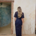 A woman with long, wavy blonde hair stands facing a textured wall and an open doorway, wearing a dark blue, floor-length gown with a subtle pattern. The setting appears to be a rustic room with sandy ground and sparse, dried grass—a perfect scene you might find at DANU PO2466. Angels Formal Wear