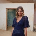 A woman stands indoors wearing a dark blue DANU PO2466 with intricate patterns from an Online Dress Shop in Australia. She has long, wavy hair and smiles gently at the camera. The background features a partially open doorway, light-colored walls, and a sandy floor. Angels Formal Wear