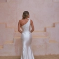 A woman with long blonde hair stands with her back to the camera, wearing a sleek, satin, one-shoulder wedding gown with a fitted silhouette and a subtle train. She is positioned in front of a textured, blush-colored backdrop that resembles a tiered wall—courtesy of SHANNON PO24100. Angels Formal Wear