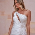 A woman with long, wavy blonde hair is wearing a white, one-shoulder gown with a cutout and elegant beading from SHANNON PO24100. She stands against a soft beige background with steps, gazing to the side. Angels Formal Wear