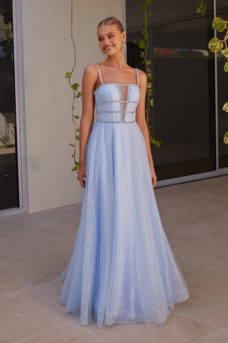 A young woman is standing outdoors, smiling and wearing a light blue, sleeveless GUINEVERE PO2473 with a flowing skirt and sparkly details from an online dress shop in Australia. The dress features thin straps and a semi-sheer panel at the front. She is standing on a tile floor with a beige wall and green vines behind her. Angels Formal Wear