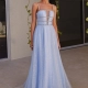 A young woman is standing outdoors, smiling and wearing a light blue, sleeveless GUINEVERE PO2473 with a flowing skirt and sparkly details from an online dress shop in Australia. The dress features thin straps and a semi-sheer panel at the front. She is standing on a tile floor with a beige wall and green vines behind her. Angels Formal Wear