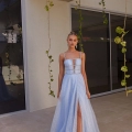 A woman stands outdoors on a patio in front of a modern building with large glass windows. She is wearing a light blue, floor-length gown with a thigh-high slit and spaghetti straps. The gown features a fitted bodice with intricate detailing, available as the GUINEVERE PO2473 at Online Dress Shop Australia. Hanging plants are seen around her. Angels Formal Wear