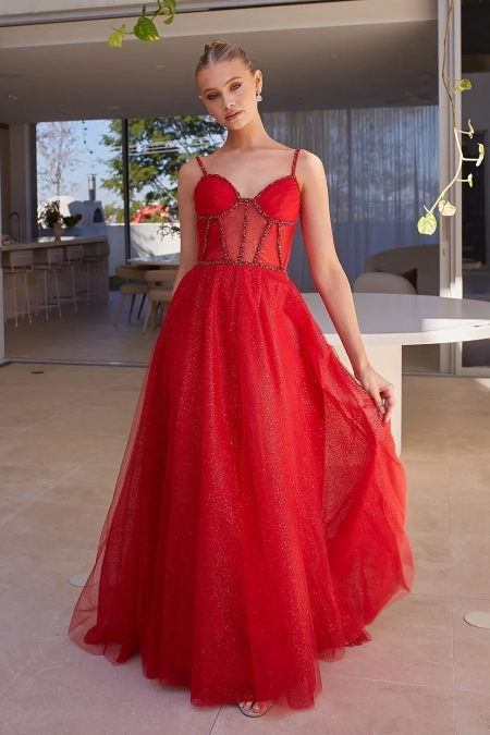 A woman stands in a modern, minimalist room. She is wearing a glamorous, floor-length red JENNA PO2474 gown with thin straps and a semi-sheer bodice adorned with glitter from an Online Dress Shop in Australia. Her hair is styled in an elegant updo as she holds a part of the dress with one hand. Angels Formal Wear