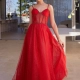 A woman stands in a modern, minimalist room. She is wearing a glamorous, floor-length red JENNA PO2474 gown with thin straps and a semi-sheer bodice adorned with glitter from an Online Dress Shop in Australia. Her hair is styled in an elegant updo as she holds a part of the dress with one hand. Angels Formal Wear