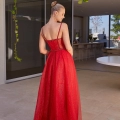 A woman with blonde hair in a bun is standing with her back to the camera, wearing JENNA PO2474 from an online dress shop in Australia. She is in a modern, open space with floor-to-ceiling windows, hanging plants, and a table with stools. The room appears bright and airy. Angels Formal Wear