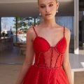 A woman with blonde hair is wearing a red dress with intricate beadwork and a see-through bodice. She is standing indoors in front of a room that opens to an outdoor patio. The background features modern furniture and greenery, showcasing the elegance available at JENNA PO2474. Angels Formal Wear