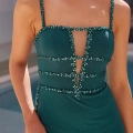 A woman is wearing a MAXWELL PO2475, a form-fitting, emerald green dress with thin straps. The dress, available at an online dress shop in Australia, features intricate bead embellishments along the straps, neckline, and four horizontal bands creating geometric patterns on the bodice. She is standing near a pool. Angels Formal Wear