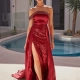 A woman stands by a pool wearing a red, strapless evening gown from ALUN PO2476, showcasing an asymmetrical off-the-shoulder design. The dress features an embellished bodice and a high slit, revealing matching high heels. The background includes outdoor architecture and a potted plant. Angels Formal Wear