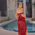 A woman stands beside a pool in an outdoor patio area, wearing a strapless, deep red dress from ALUN PO2476 with intricate gold detailing. Her hair is styled in an elegant updo, and she poses with one leg slightly forward, gazing over her shoulder towards the camera. Angels Formal Wear