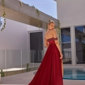 A woman in a strapless, flowing red gown from ALUN PO2476 stands by a modern outdoor pool area. Her dress features a long, elegant train. She has her hair styled in a bun and is looking over her shoulder. The background includes a building with tall windows and trailing green plants. Angels Formal Wear