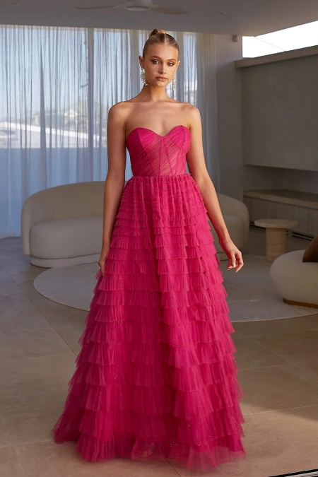 A person stands in a modern room wearing a strapless, floor-length fuchsia gown from DERYA PO2481, featuring a fitted bodice and layered ruffle skirt. Their hair is styled up, and they accessorize with dangling earrings. The background features beige furnishings and sheer white curtains. Angels Formal Wear