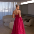 A woman with blonde hair styled in an elegant updo is wearing a strapless, layered magenta DERYA PO2481 from Angels Formal Wear. She is standing in a modern, minimalist living room with white furniture, gazing out a large window with sheer curtains. Angels Formal Wear