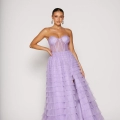 A woman in a strapless, lavender, tiered tulle gown from Angels Formal Wear poses against a plain white background. The DERYA PO2481 creation features a structured bodice and a high slit in the skirt. She wears her hair pulled back and has statement earrings. Angels Formal Wear