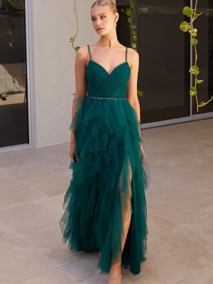 A woman in an elegant green, ruffled evening gown with sheer overlay and thin straps stands on a light-colored floor. She is wearing strappy sandals and has her hair styled in a sleek updo. The background features a modern wall with hanging plants, adding to the chic SAGA PO2483 of her look. Angels Formal Wear