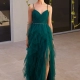 A woman in an elegant green, ruffled evening gown with sheer overlay and thin straps stands on a light-colored floor. She is wearing strappy sandals and has her hair styled in a sleek updo. The background features a modern wall with hanging plants, adding to the chic SAGA PO2483 of her look. Angels Formal Wear