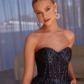 A woman wearing an elegant strapless black gown adorned with intricate lace and sequins. She has blonde hair styled in a sleek bun and accessorizes with statement dangling earrings. Standing against a backdrop featuring sheer curtains and modern furniture, she epitomizes the sophisticated charm of EYRA PO2491. Angels Formal Wear