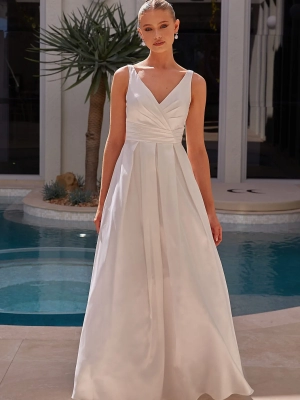 A person stands in front of a pool wearing a sleeveless white gown from YARELI PO24110, featuring a fitted bodice and a V-neckline. The gown flows into an elegant skirt. The background showcases a modern building facade and a potted plant. The person has neatly styled hair and wears earrings. Angels Formal Wear