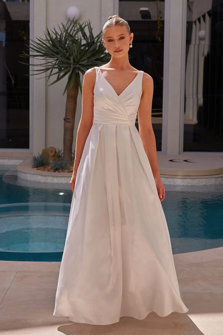 A person stands in front of a pool wearing a sleeveless white gown from YARELI PO24110, featuring a fitted bodice and a V-neckline. The gown flows into an elegant skirt. The background showcases a modern building facade and a potted plant. The person has neatly styled hair and wears earrings. Angels Formal Wear