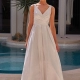 A person stands in front of a pool wearing a sleeveless white gown from YARELI PO24110, featuring a fitted bodice and a V-neckline. The gown flows into an elegant skirt. The background showcases a modern building facade and a potted plant. The person has neatly styled hair and wears earrings. Angels Formal Wear