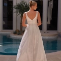 A woman stands with her back to the camera wearing an elegant, sleeveless white wedding dress from YARELI PO24110, featuring a V-back. Her hair is styled in a neat bun as she poses in front of a luxurious pool, surrounded by modern architecture, including large windows and a palm tree. Angels Formal Wear