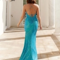 A woman with wavy brown hair is wearing a sparkling blue, floor-length gown with an open back and crossing straps. She is walking on a sunlit patio with white columns and decorative wall lamps, towards a glass-paned door, embodying the elegance you'd find at the Sunshine Coast Dress Shop's NEVA PO964. Angels Formal Wear