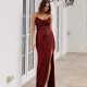 A woman stands in front of a white building with arched windows, wearing a sleeveless, deep red sequin gown with a high slit from DIVA PO959. She has shoulder-length brown hair and wears nude heels. The ambiance is elegant, with ornate wall-mounted lanterns. Angels Formal Wear