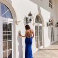 A woman wearing a blue, backless evening gown stands with her back to the camera, facing a white, arched wall adorned with black, decorative lanterns. Sunlight casts a shadow of her form onto the wall, and tall windows with white frames are set beside her. It's an elegant scene reminiscent of a chic TALIA PO963 on Sunshine Coast. Angels Formal Wear