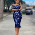 A woman stands confidently on a sidewalk, wearing an elegant off-shoulder navy blue dress with a floral pattern. She has curly hair, hoop earrings, and is wearing high-heeled sandals. The background shows an urban street scene with trees and buildings. ADRIANNA JX4066 absolutely stuns in this look. Angels Formal Wear