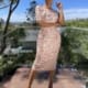 A woman stands outdoors on a wooden deck, wearing a fitted, light pink lace dress from ADRIANNA JX4066 and open-toe heels. She poses with one hand on her hip and another touching her face. The background consists of a lush garden and trees against a clear blue sky. Angels Formal Wear