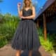 A woman in a black, sleeveless, knee-length dress stands on a wooden pathway surrounded by lush greenery. With long, blonde hair and silver high heels (ASTRA JX5030), she gazes up at the clear blue sky. Angels Formal Wear