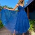 A woman in a blue, shimmery, pleated ASTRA JX5030 dress poses on a wooden path surrounded by greenery. She holds one side of her dress out, showcasing its volume. With wavy blonde hair and silver strappy heels, she stands against a backdrop featuring a building and a clear sky. Angels Formal Wear