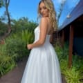 A woman with long blonde hair is standing on a wooden pathway surrounded by lush greenery. She is wearing a white, flowing dress and looking over her shoulder towards the camera with a calm expression. The clear blue sky above seems infinite, reminiscent of the serene vistas often captured by ASTRA JX5030. Angels Formal Wear