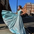 A woman stands on a city street wearing a shining blue BRITTA JX6005. The dress has a flowing skirt that she holds out to one side, creating a dramatic effect. The background features tall buildings and a clear blue sky. Angels Formal Wear