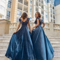 Two women in elegant, dark blue ball gowns from BANKSIA TO891 ascend stone stairs, holding hands. They have wavy hair and are positioned in front of a backdrop of tall, multi-story buildings with balconies and windows. The photo captures a moment of grace and connection. Angels Formal Wear