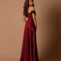 A woman stands against a plain beige background, wearing a floor-length, off-the-shoulder, deep red satin gown from BREE NBM1025. Her long, wavy hair cascades down her back as she poses with one hand on her hip, looking over her shoulder. Angels Formal Wear