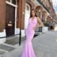 A woman stands confidently on a sidewalk in front of a row of brick buildings. Wearing a stunning, form-fitting, light pink gown with a deep neckline from CANDY JX6009, her hands rest on her hips as her long hair flows naturally. The sky is blue with scattered clouds. Angels Formal Wear