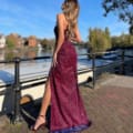 A woman wearing a sparkling maroon gown from CLARISSA JP136 poses with her back to the camera by a waterfront. The gown has a high slit that reveals her legs, and she is wearing high heels. The background features boats, a paved walkway, and trees under a clear blue sky. Angels Formal Wear