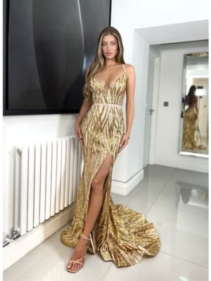 A woman stands indoors wearing a sparkling gold gown from CHICA JX6024, featuring a deep V-neck and thigh-high slit. She has long, wavy hair and is posing with one hand on her hip in a modern, minimalistic room with a large mirror and radiator in the background. Angels Formal Wear