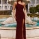 A woman stands in front of a large decorative fountain with a stately white building in the background. She is wearing a sleeveless, one-shoulder dark red gown from CARMEN PO966, complete with a thigh-high slit and strappy heels, striking a confident pose. Angels Formal Wear