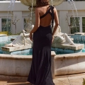 A person wearing a black, sleeveless, backless CARMEN PO966 with a long skirt stands on a paved path in front of a large ornate fountain with multiple water streams. Their back is towards the camera, and they have shoulder-length wavy hair. Angels Formal Wear