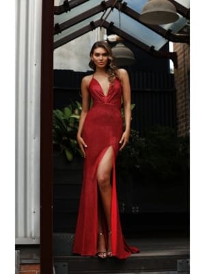 A woman in a sleeveless, deep red DAZZLING JX3082 with a plunging neckline and high slit poses outdoors under a structure with hanging lights. She has long hair styled in loose waves and is wearing high-heeled sandals, standing confidently in front of a black background with greenery. Angels Formal Wear