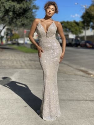 A woman stands on a street in daylight, wearing a sparkling, form-fitting, silver gown with a plunging neckline and thin straps from DAZZLE JP111. She has curly hair and a confident pose, with hands resting on her hips. Trees, buildings, and parked cars are visible in the background. Angels Formal Wear